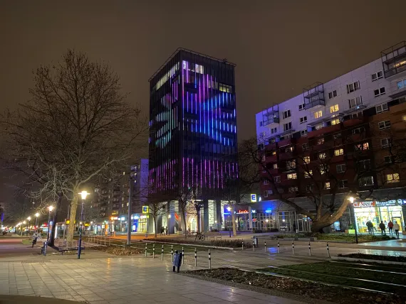 Häuserzeile an der Straße ein Hochhaus sticht beleuchtet hervor. Es ist abends. 